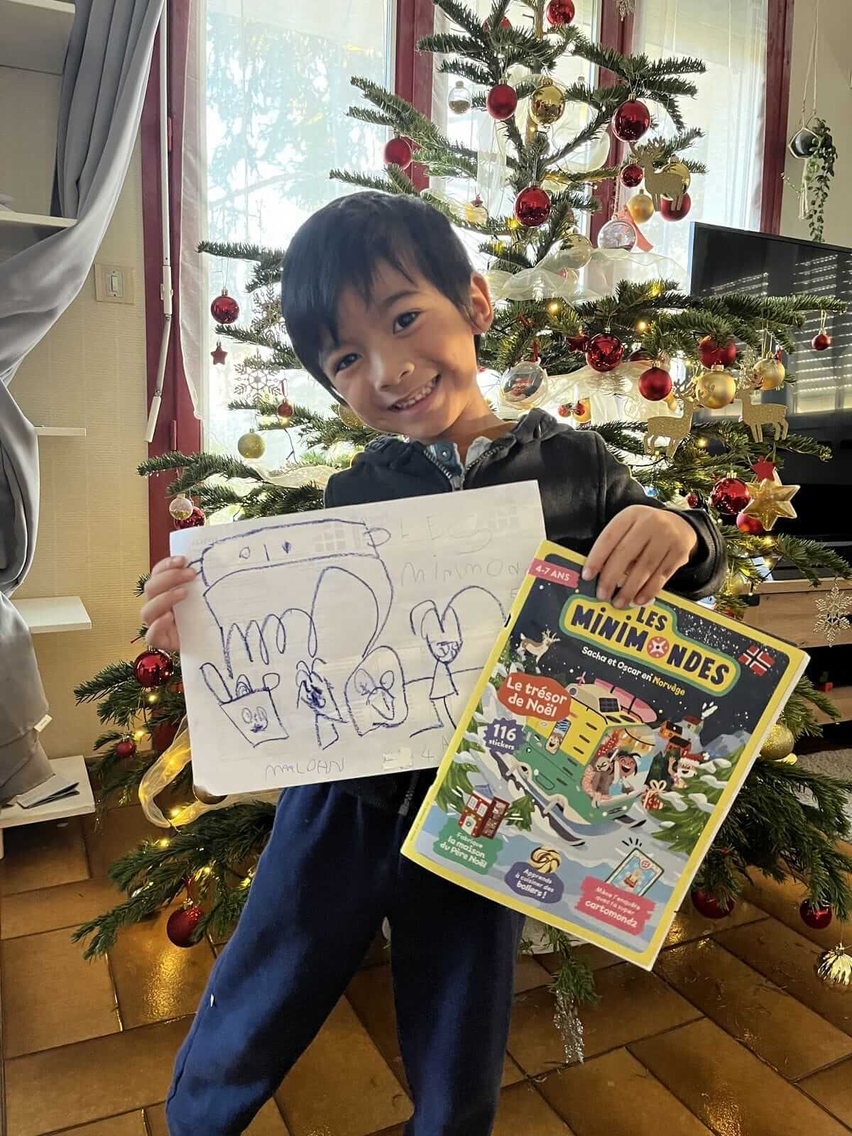Un enfant souriant abonné au magazine Les Mini Mondes posant fièrement devant son sapin de Noël avec un dessin.