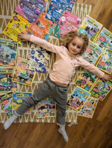 Une petite fille allongée sur le sol, entourée de magazines éducatifs, les bras ouverts comme pour embrasser son univers de lecture.