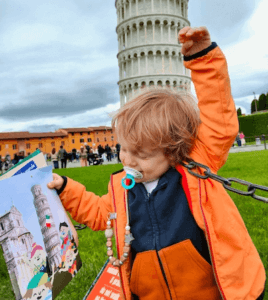 Un enfant découvre le monde à travers un magazine éducatif devant la Tour de Pise, alliant apprentissage et voyage.