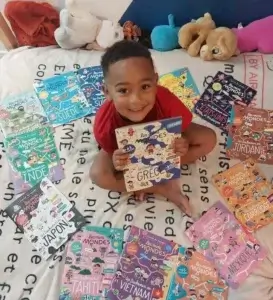 Petit garçon souriant assis sur un lit, tenant un magazine Les Mini Mondes sur la Grèce, entouré de plusieurs autres numéros dédiés à différents pays